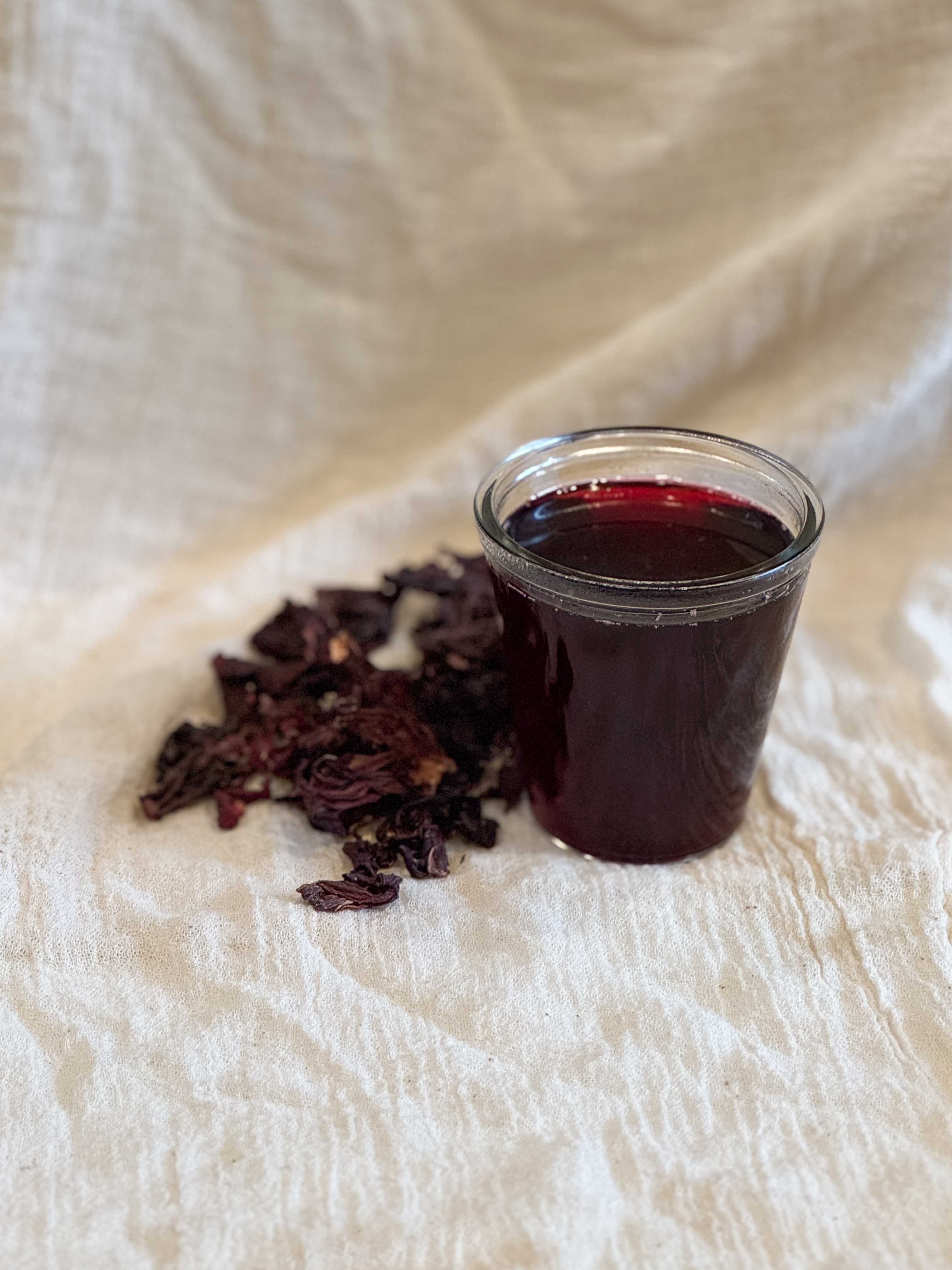 A glass jar of jamaica simple syrup on a white muslin cloth next to dried hibiscus flowers.