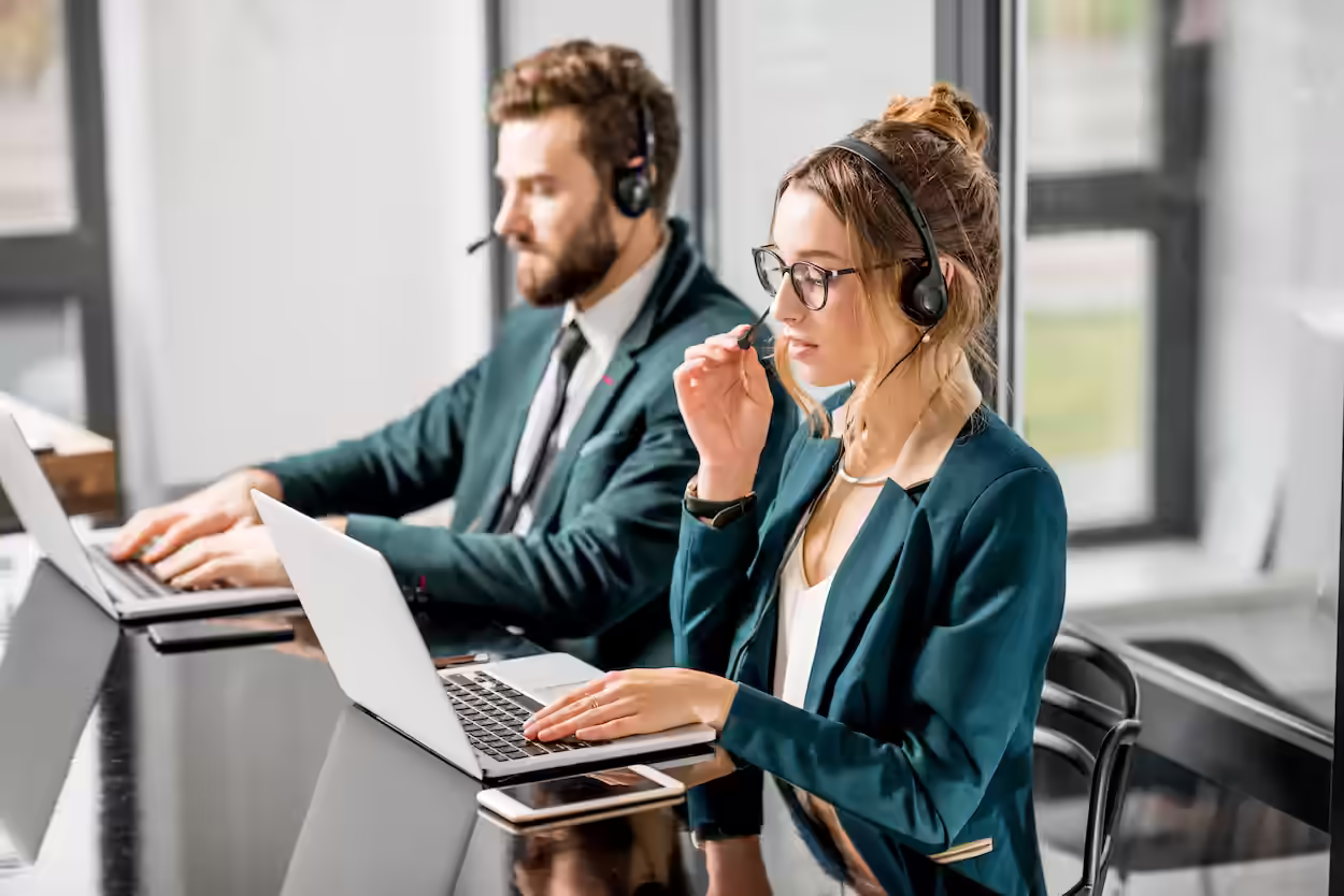 Two customer service representatives wearing headsets and working on laptops in a modern office.