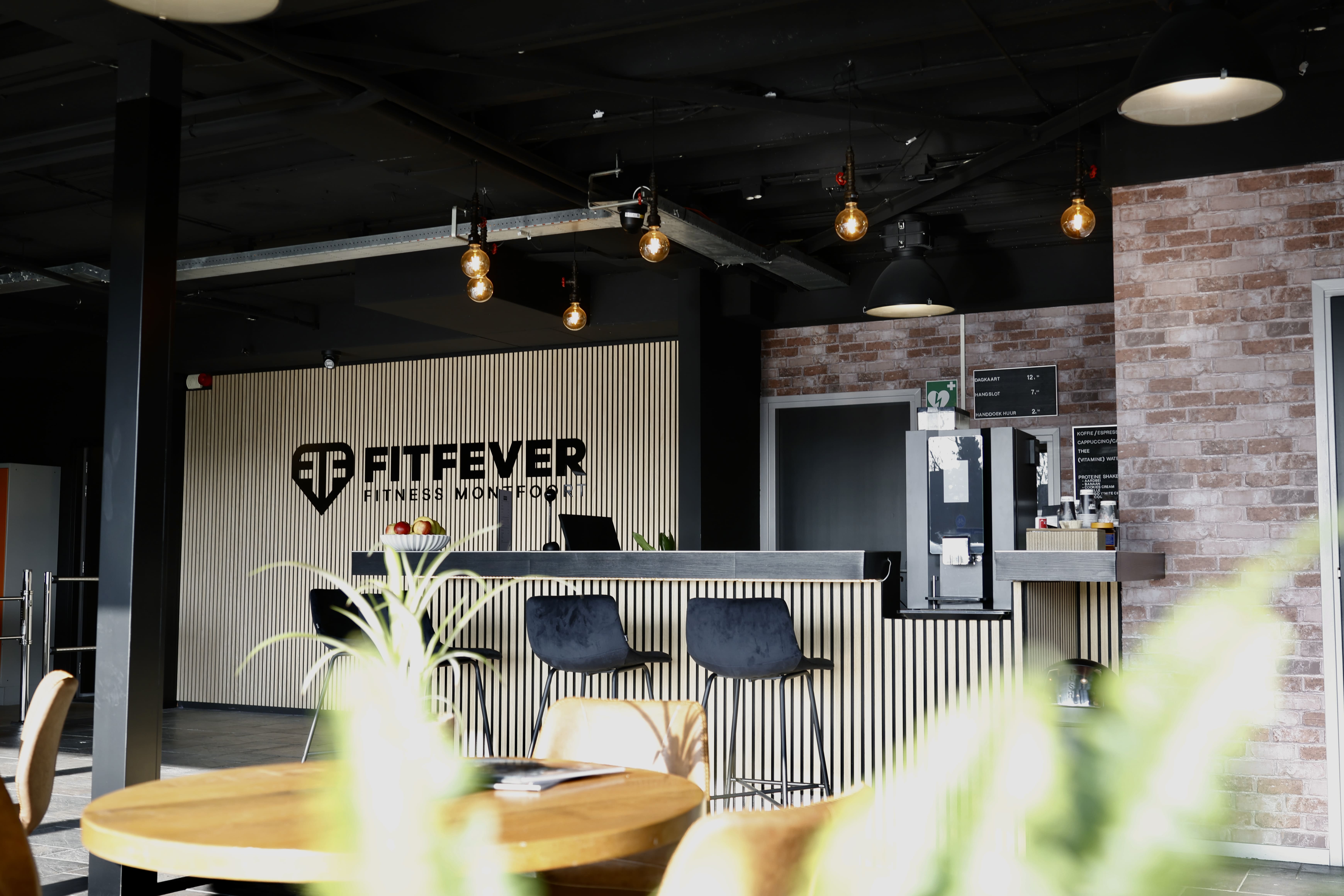 Modern gym reception area with black bar stools, a coffee machine, hanging light bulbs, and the FitFever logo on a striped wall.