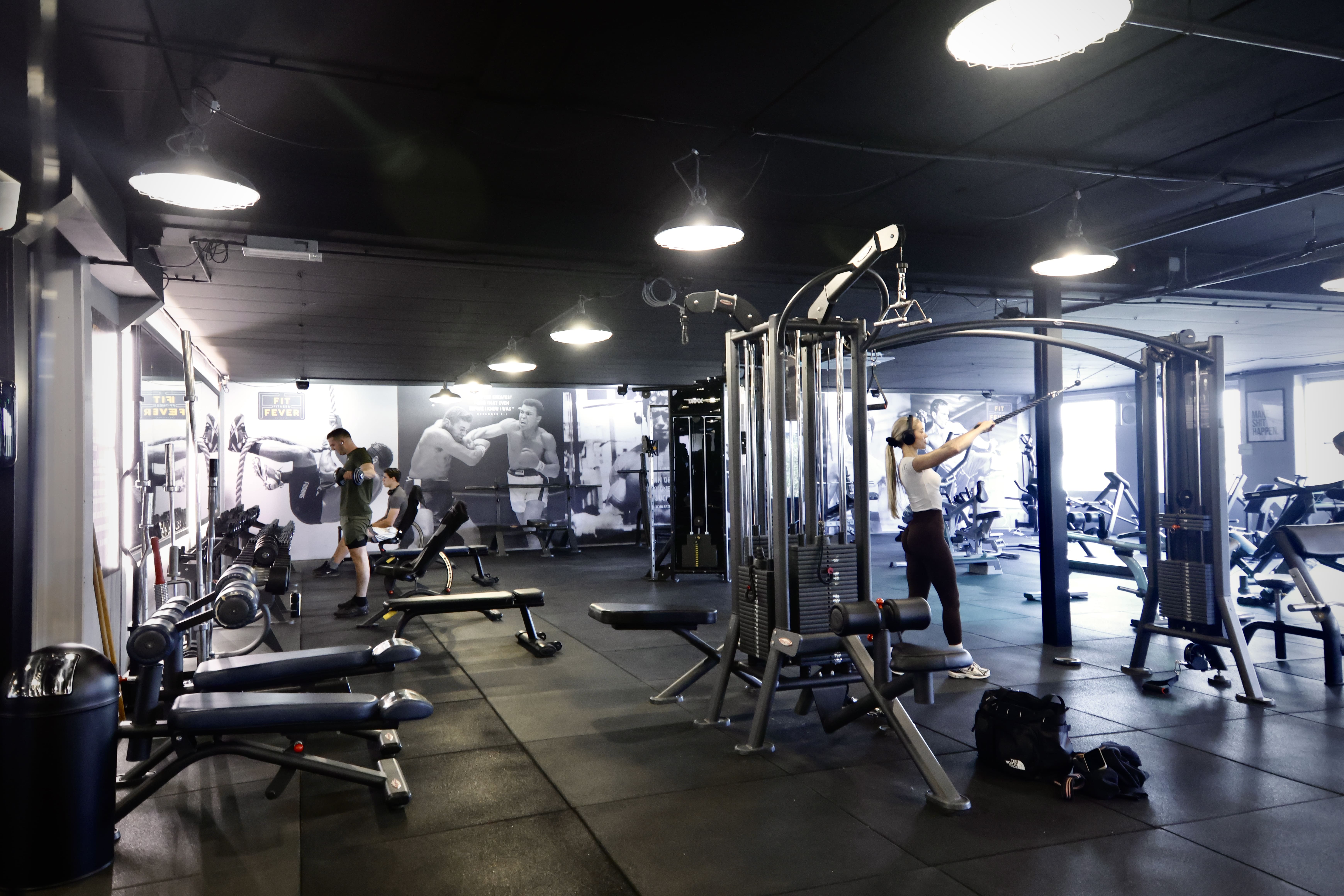 Interior of a gym with people working out using various exercise machines and free weights, featuring wall posters of boxing scenes.