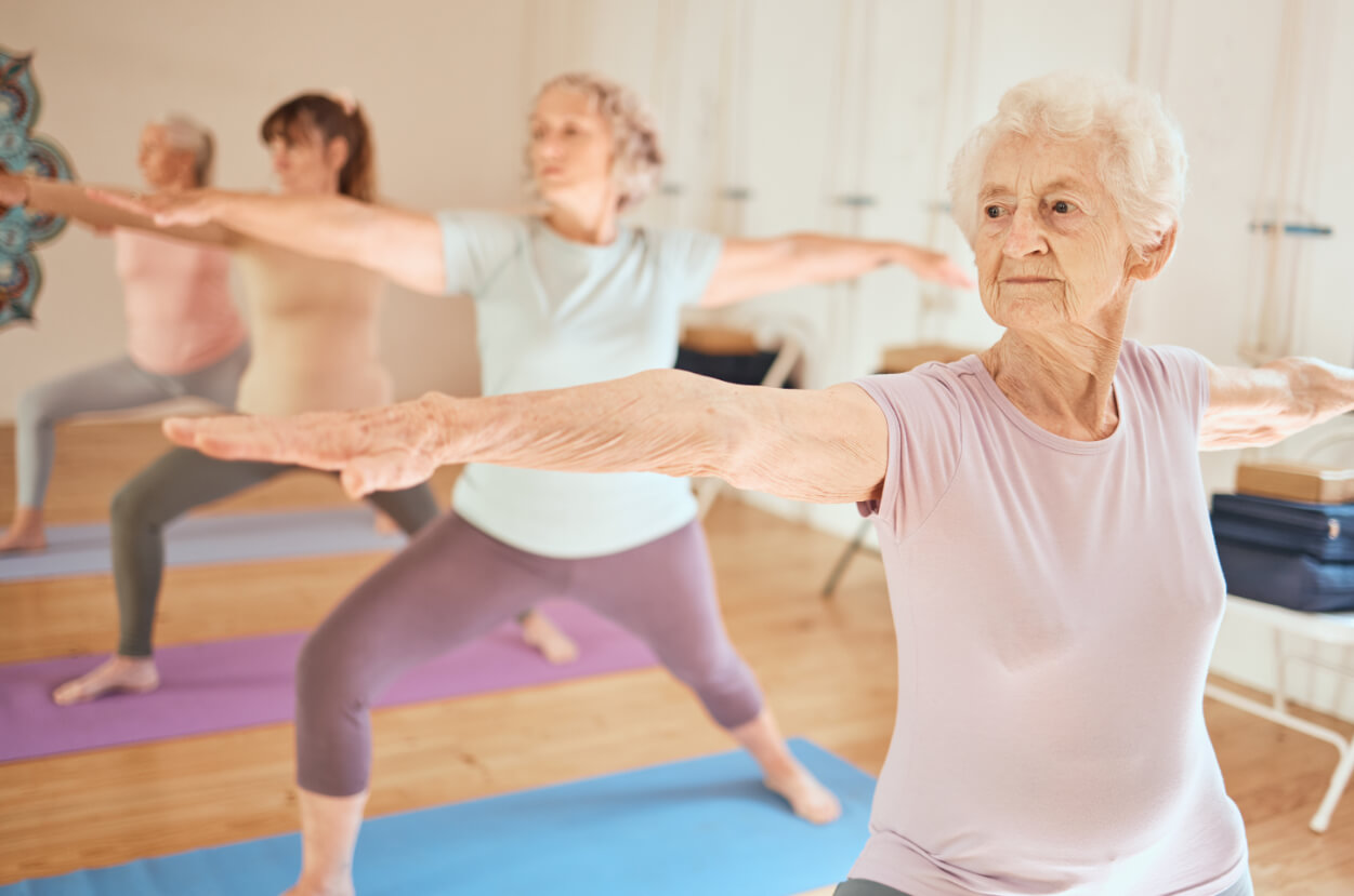Four elderly women practicing yoga with arms extended in a bright studio on colorful mats.