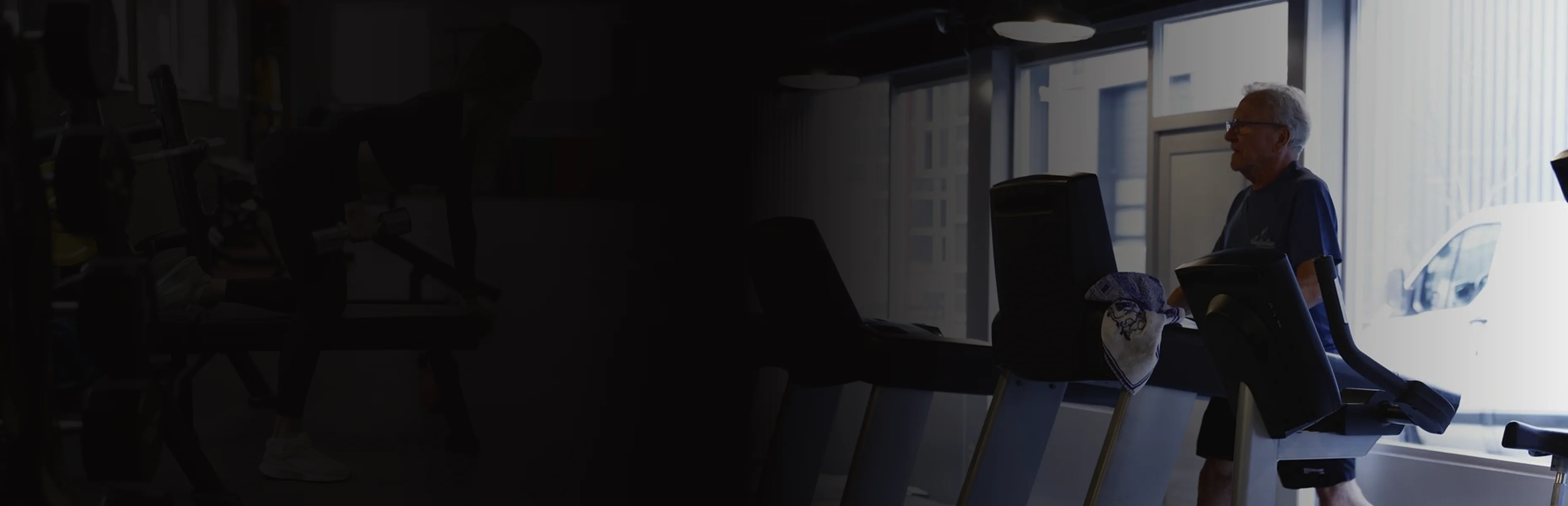 Elderly man walking on a treadmill near large windows in a gym.