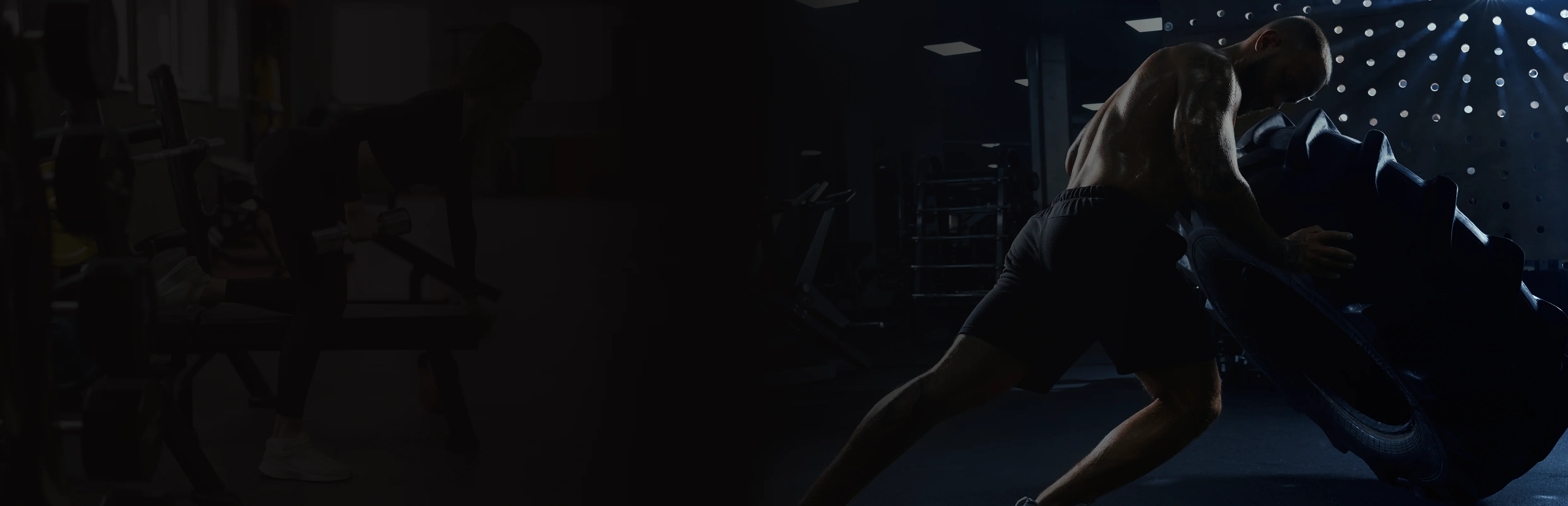 Muscular man in shorts pushing a large tire in a dimly lit gym.