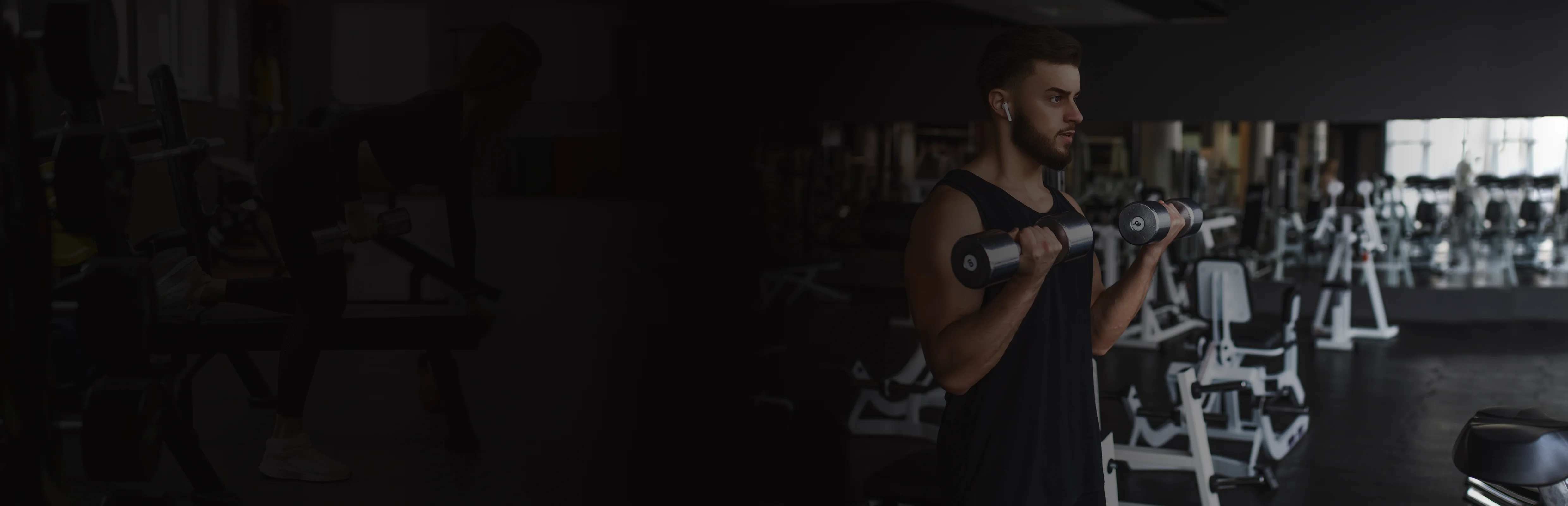 Man in black tank top lifting dumbbells in a gym filled with exercise machines.