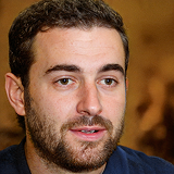 Portrait of a man with short curly hair, beard, and mustache speaking in front of a blurred brown background.