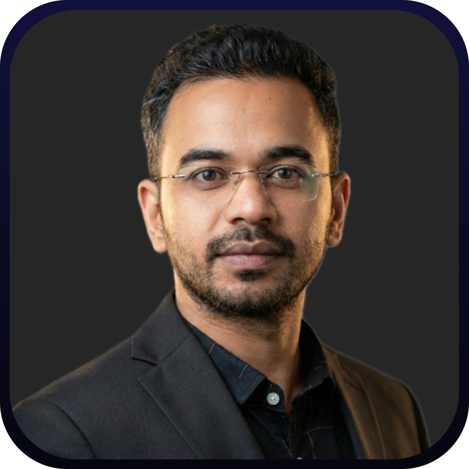 Portrait of a man with short dark hair and glasses wearing a black suit jacket and dark shirt against a dark background.