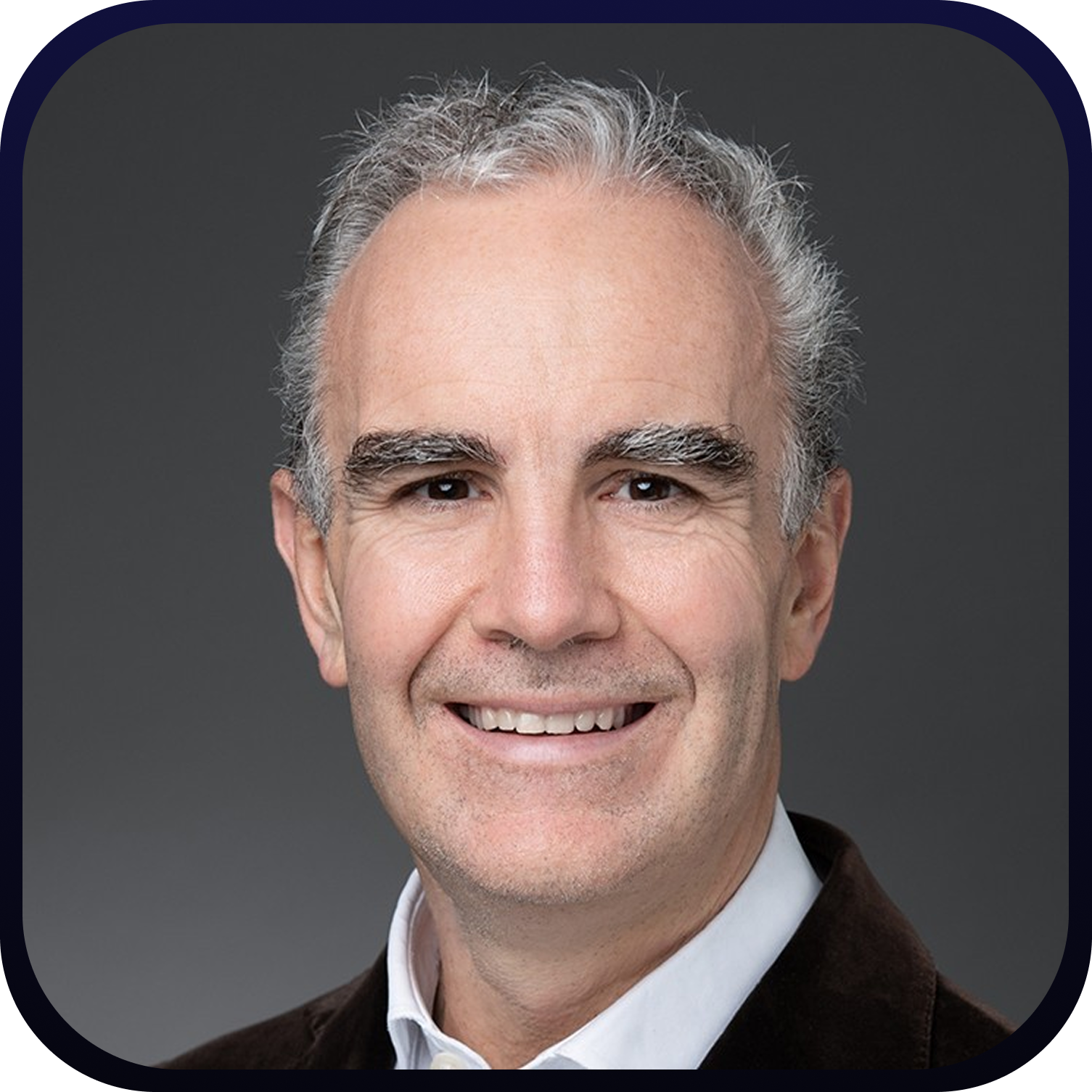 Smiling middle-aged man with gray hair, wearing a white shirt and dark blazer against a gray background.