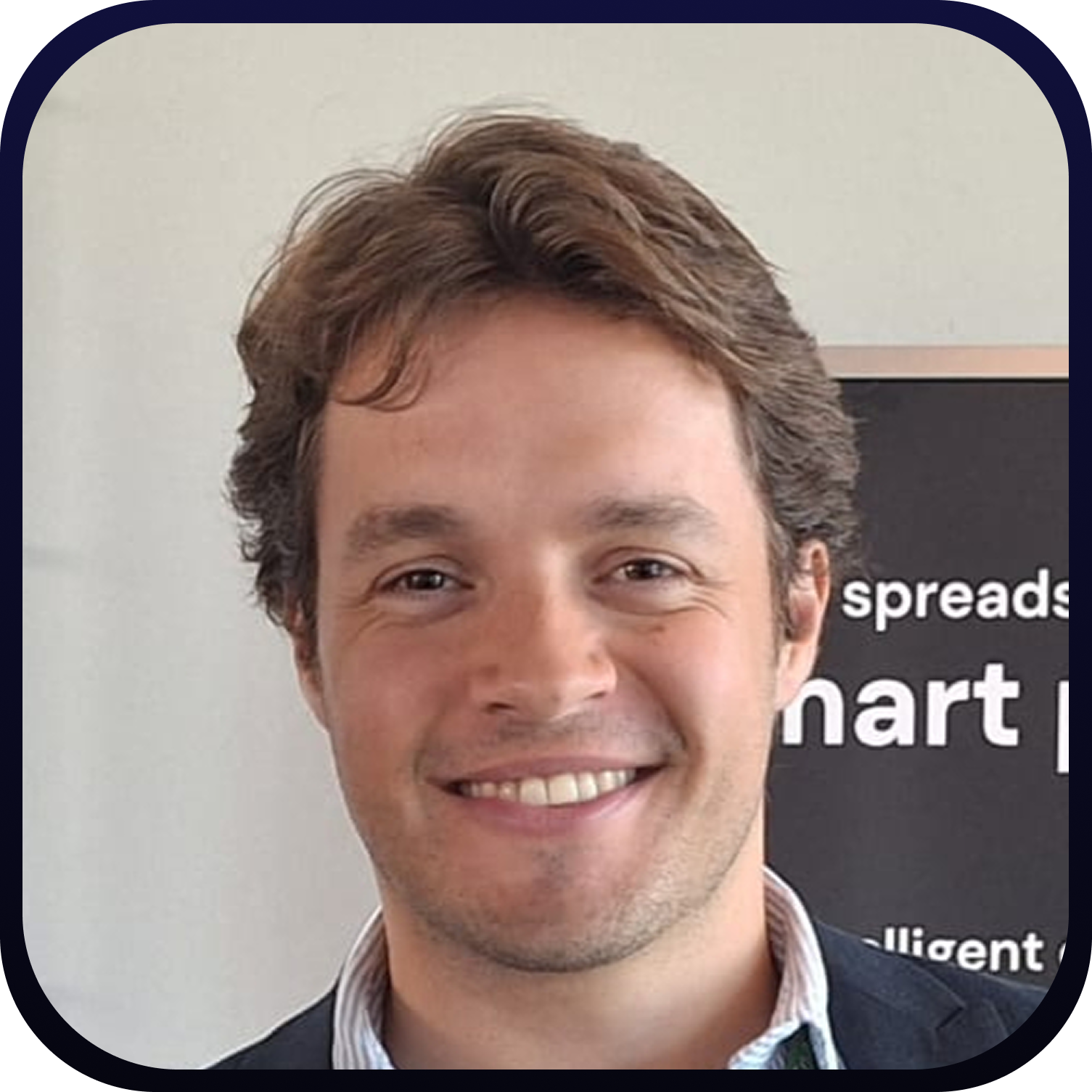 Smiling man with short brown hair wearing a dark blazer and light shirt against a light background with partial text.