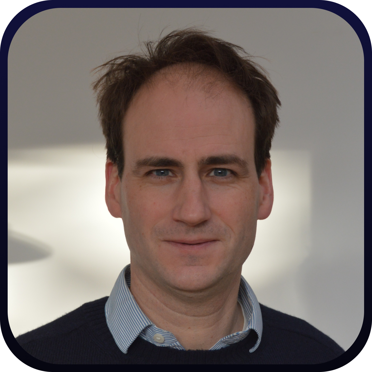 Smiling man with short brown hair wearing a dark blazer and light shirt against a light background with partial text.