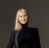 Smiling woman with long blonde hair wearing a black turtleneck against a dark background.