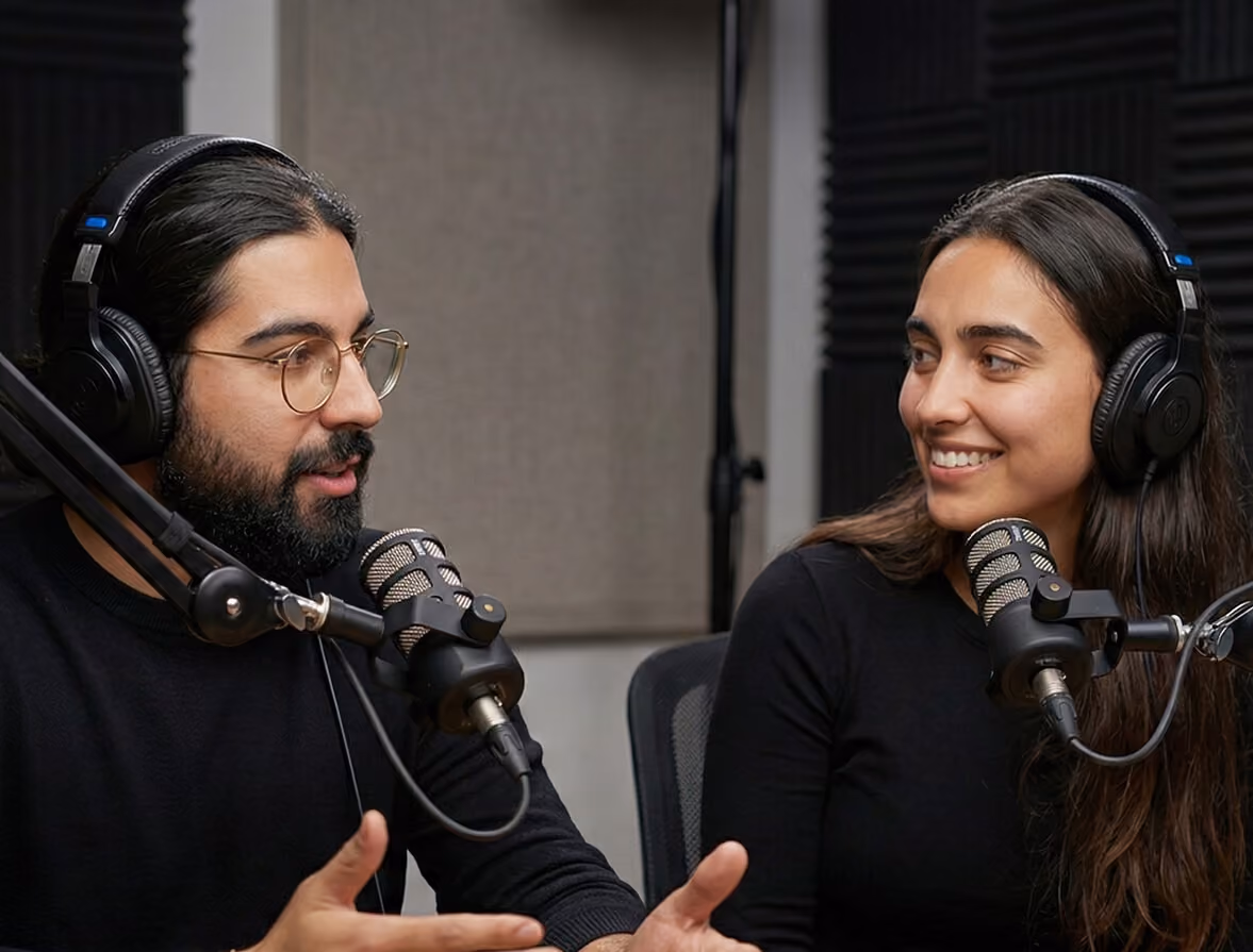 Two people wearing headphones speaking into microphones in a recording studio.
