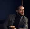 Bearded man in a gray blazer and black turtleneck sitting and looking thoughtfully to the side against a dark blue background.
