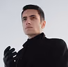 Young man wearing black gloves and a black turtleneck looking confidently into the distance against a clear sky.