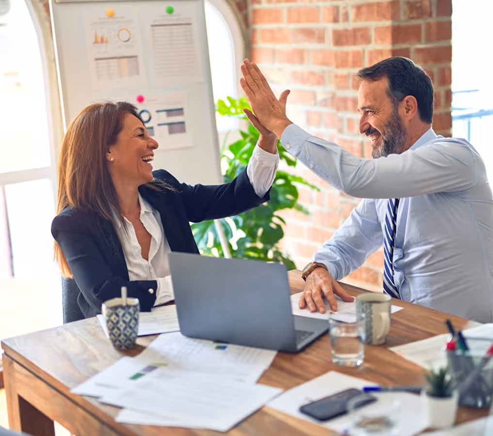 Two business colleagues smiling and giving each other a high five at a desk with laptop and documents.