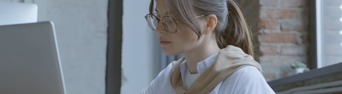 Young woman with glasses working on a laptop in a room with a brick wall and window.
