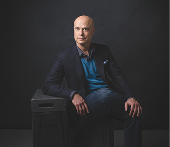 Ben Morley in a navy blazer, blue sweater, and jeans sitting on a black stool against a dark background.