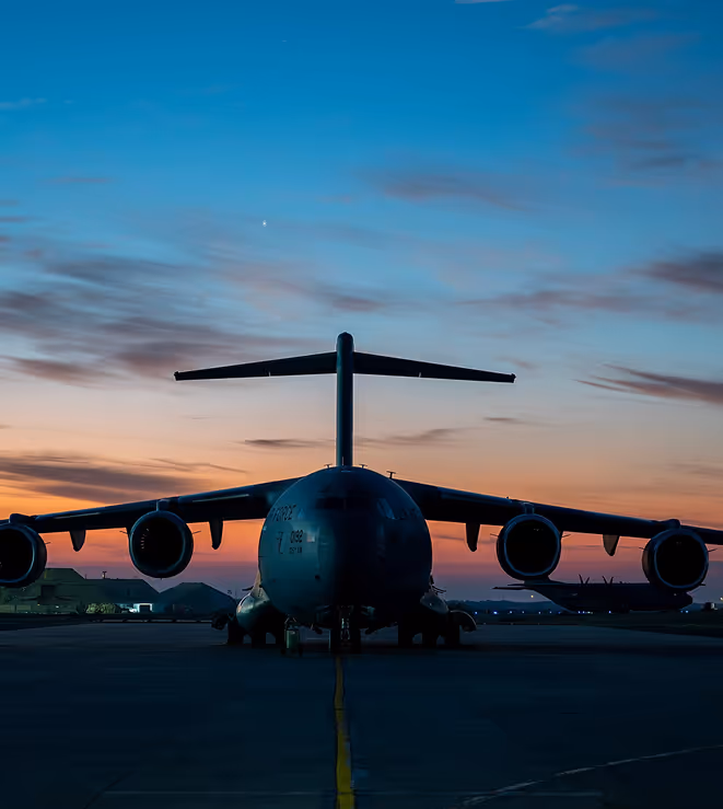 Front view of a large military cargo airplane on a runway during sunset with colorful sky.