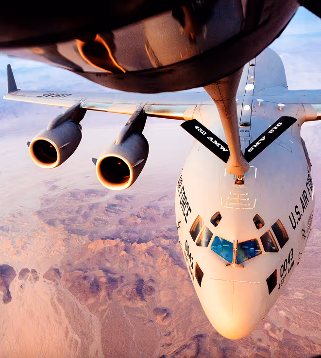 Refueling boom connected to a U.S. Air Force tanker aircraft flying above a desert landscape during sunset.