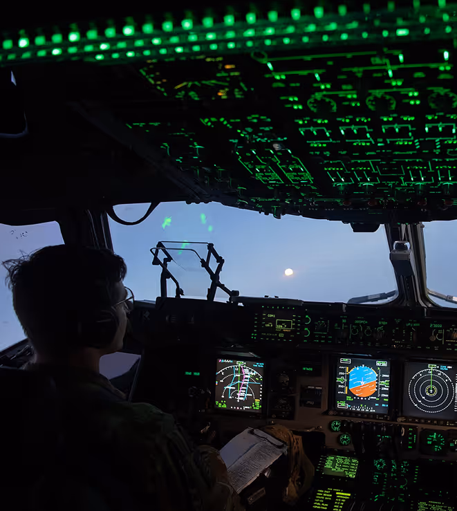 Pilot seated in a dimly lit cockpit with illuminated green controls and flight instruments visible at dusk.