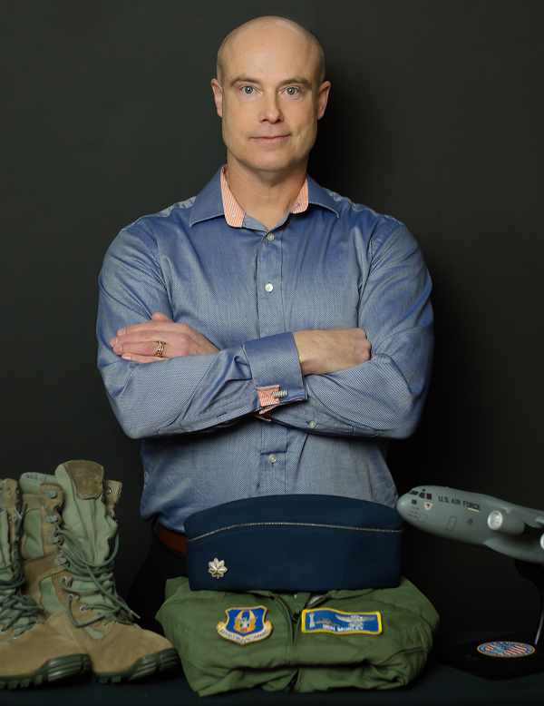 Ben Morley in a blue shirt standing with arms crossed behind military boots, a flight uniform, a blue cap with a silver leaf insignia, and a model U.S. Air Force airplane on a black background.