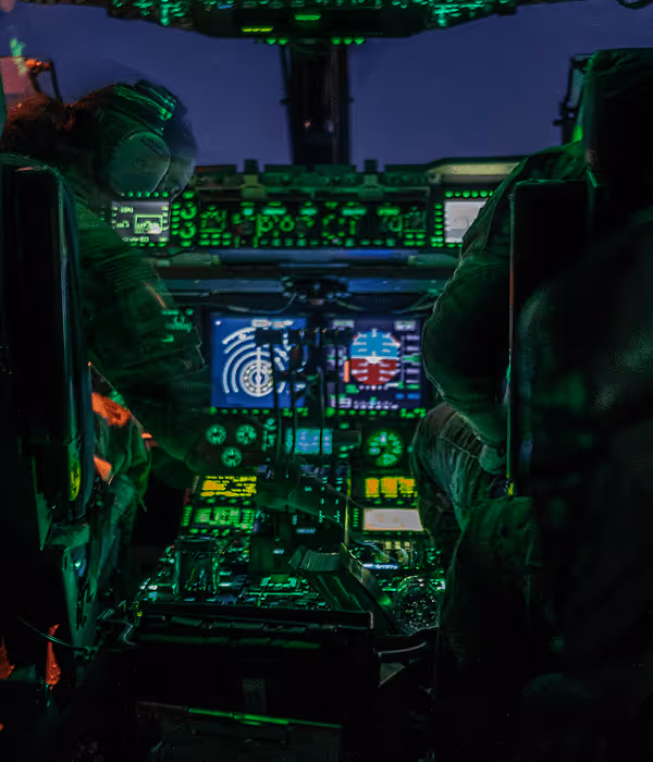 Two pilots in a dimly lit cockpit with illuminated instrument panels and flight controls.