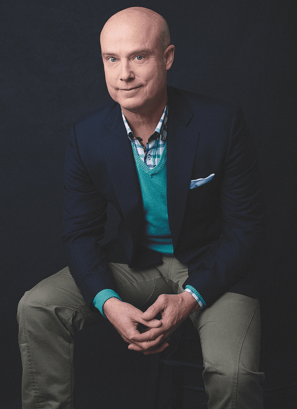 Ben Morley wearing a navy blazer, turquoise sweater vest, checkered shirt, and beige pants sitting with hands clasped.