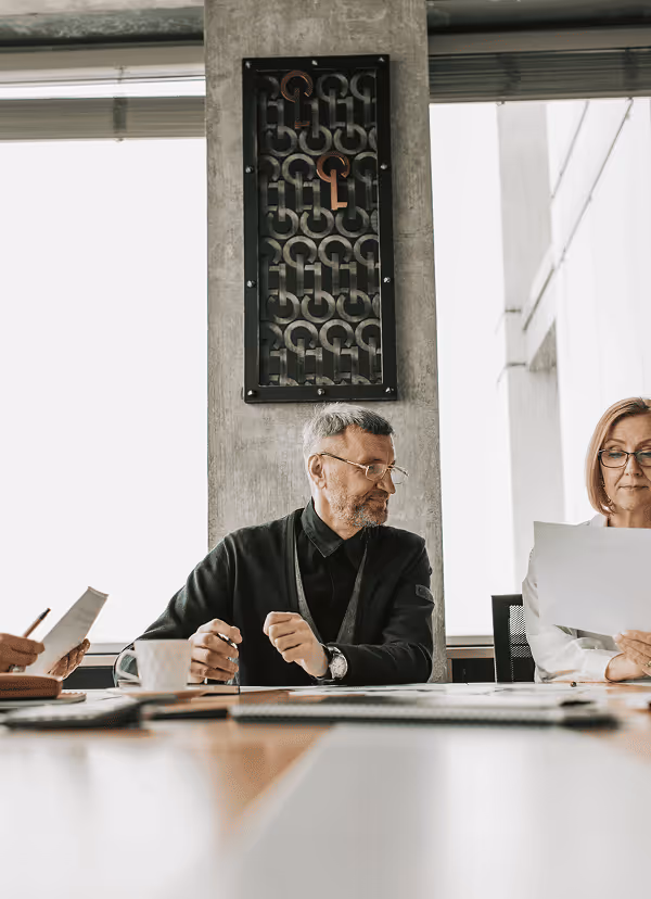 Two professionals sitting at a conference table reviewing documents in a modern office setting with large windows and a decorative wall panel.
