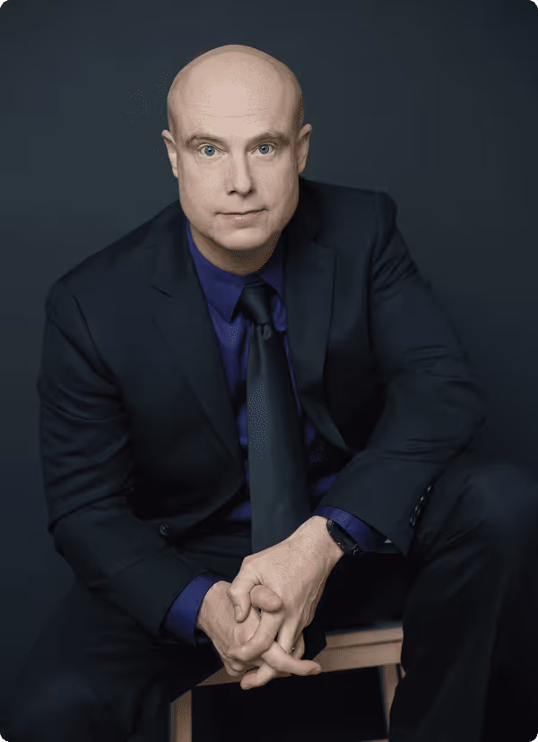 Ben Morley in a dark suit with a purple shirt and black tie sitting on a wooden stool against a dark background.