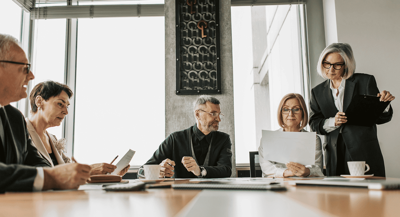 Five business professionals in a meeting room reviewing documents together around a conference table.