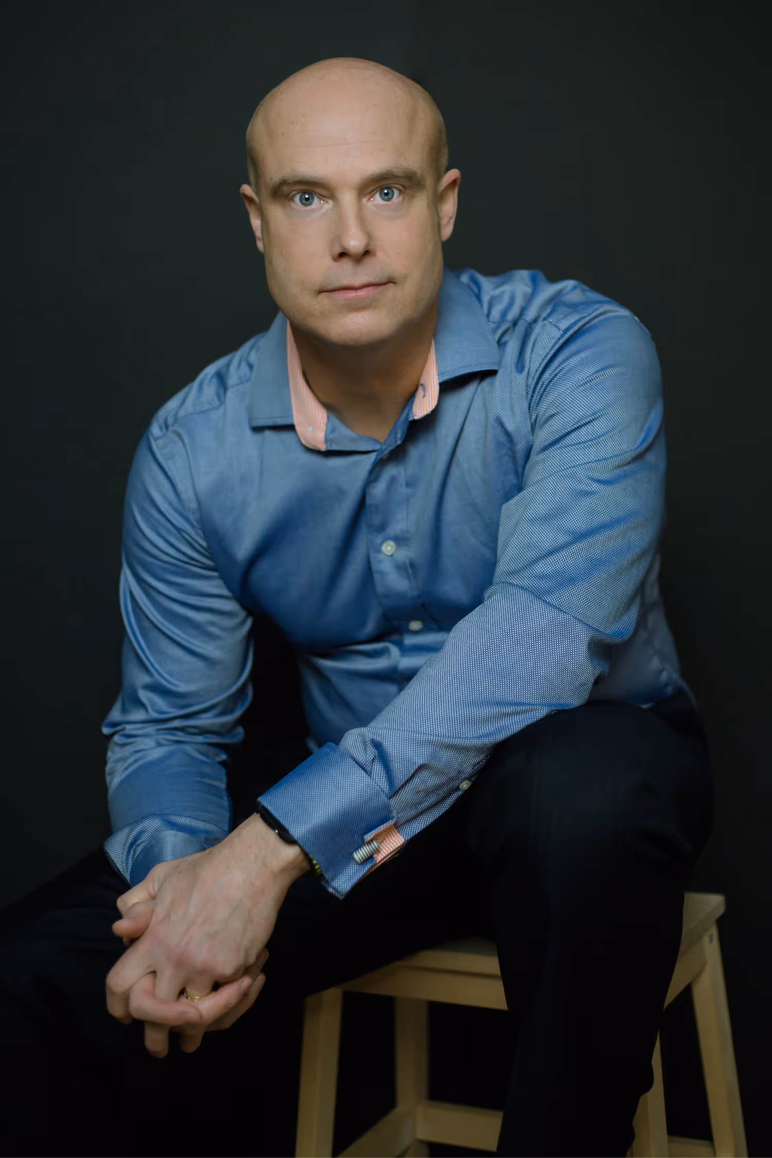 Ben Morley wearing a blue dress shirt sitting on a wooden stool against a dark background.