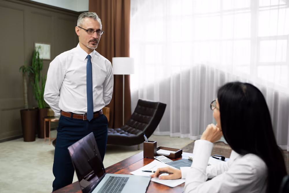 Man in white shirt and blue tie standing with hands behind back talking to seated woman with glasses in an office.
