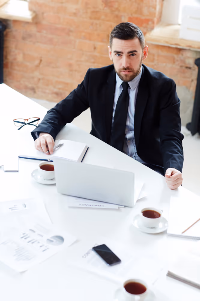 Businessman in a black suit sitting at a white desk with a laptop, coffee cups, and documents.