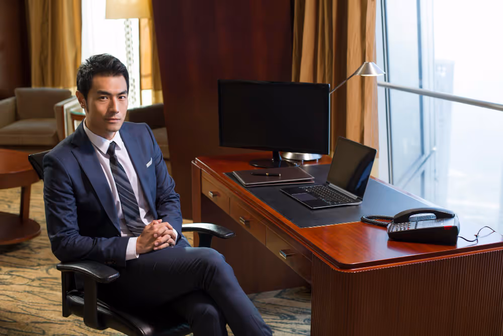 Businessman sitting in an office chair beside a wooden desk with a laptop, monitor, desk phone, and lamp, looking at the camera.