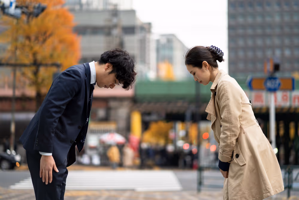 Two people bowing respectfully to each other on a city street with blurred buildings and autumn trees in the background.