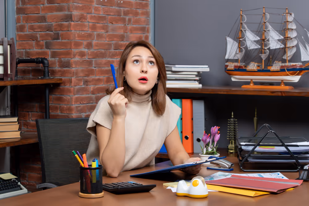 Woman sitting at a desk holding a pen, looking upwards thoughtfully with office supplies and a model ship behind her.