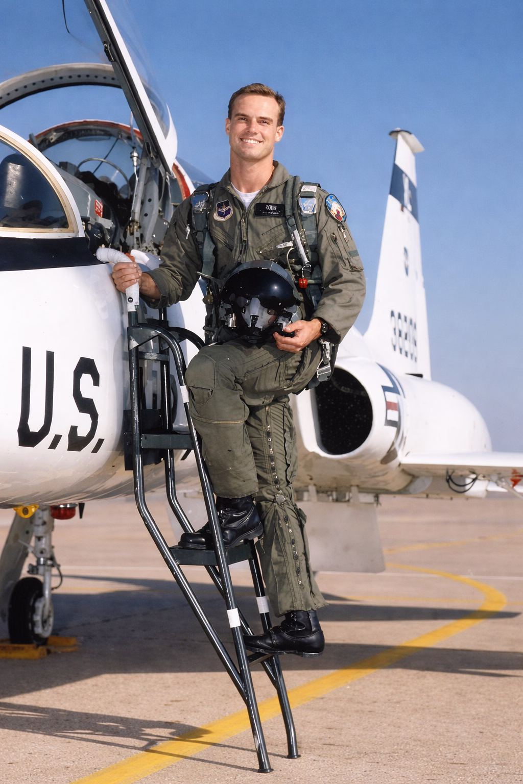 Ben Morley in green flight suit holding a helmet, standing on ladder beside a U.S. Air Force jet on runway.