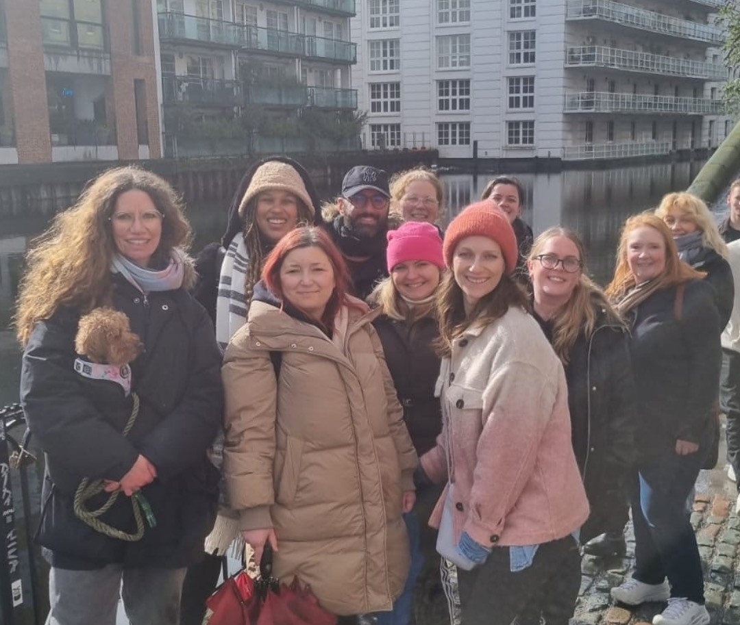 Group of people wearing winter clothes standing on a cobblestone path near a water canal with buildings in the background.