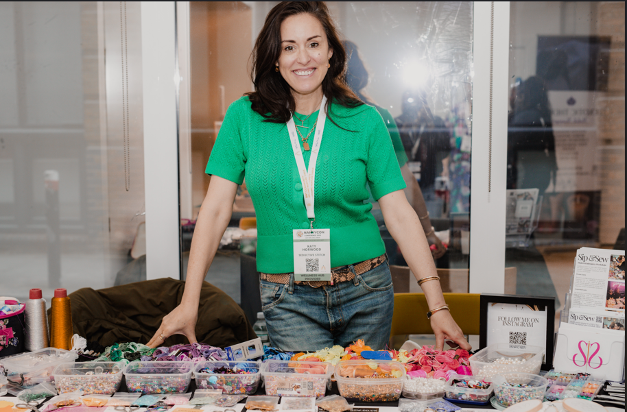 Smiling woman in a green sweater standing behind a table filled with colorful beads and crafting supplies at a vendor booth.