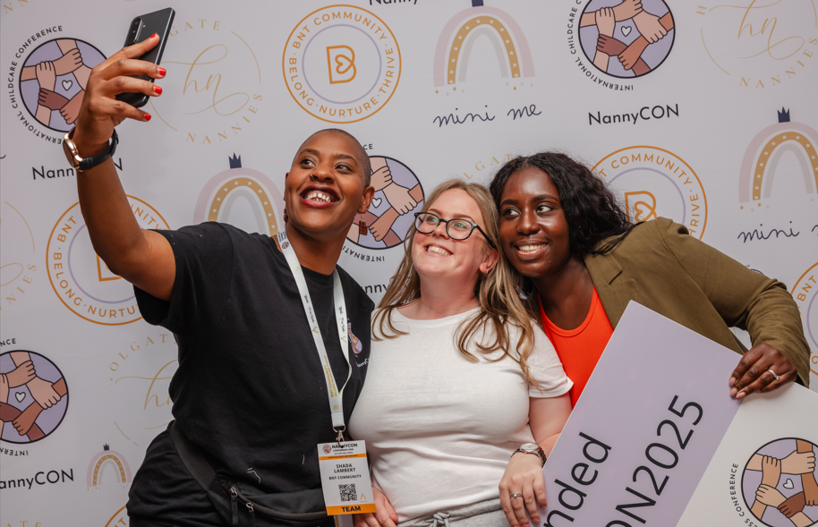 Three smiling women posing for a selfie in front of a backdrop with NannyCON and related logos, one holding a sign with #NANNYCON2025.