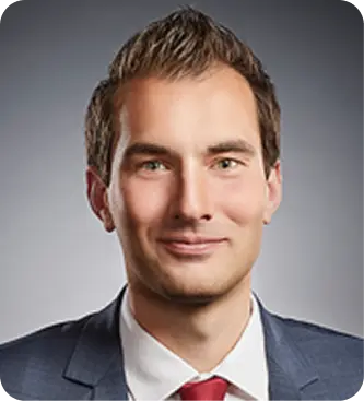 Porträt eines Mannes mit kurzem braunem Haar, dunkelblauem Anzug, weißem Hemd und roter Krawatte vor grauem Hintergrund.