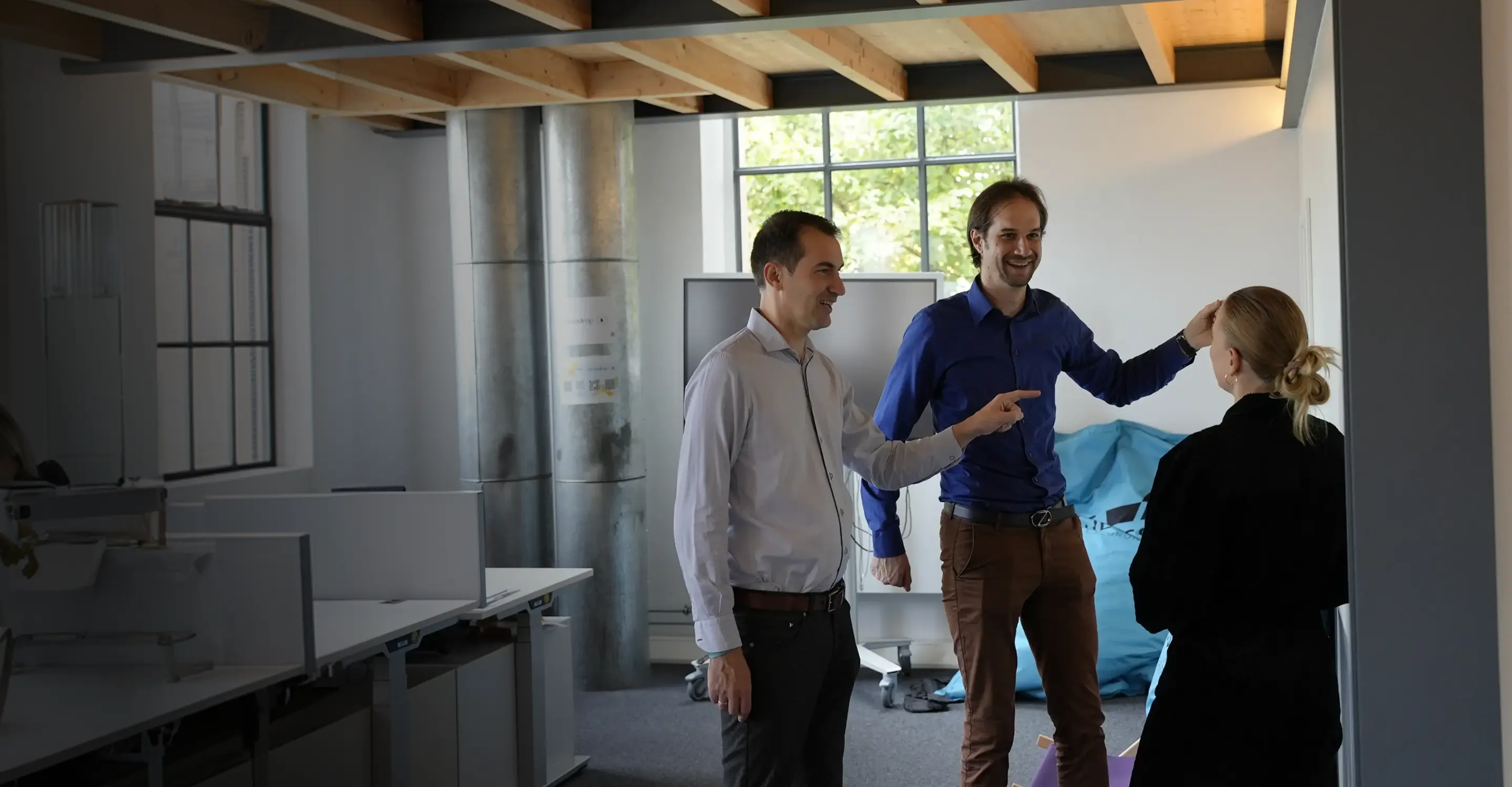 Drei Personen im Büro führen ein freundliches Gespräch, zwei Männer lächeln und zeigen auf eine Frau.