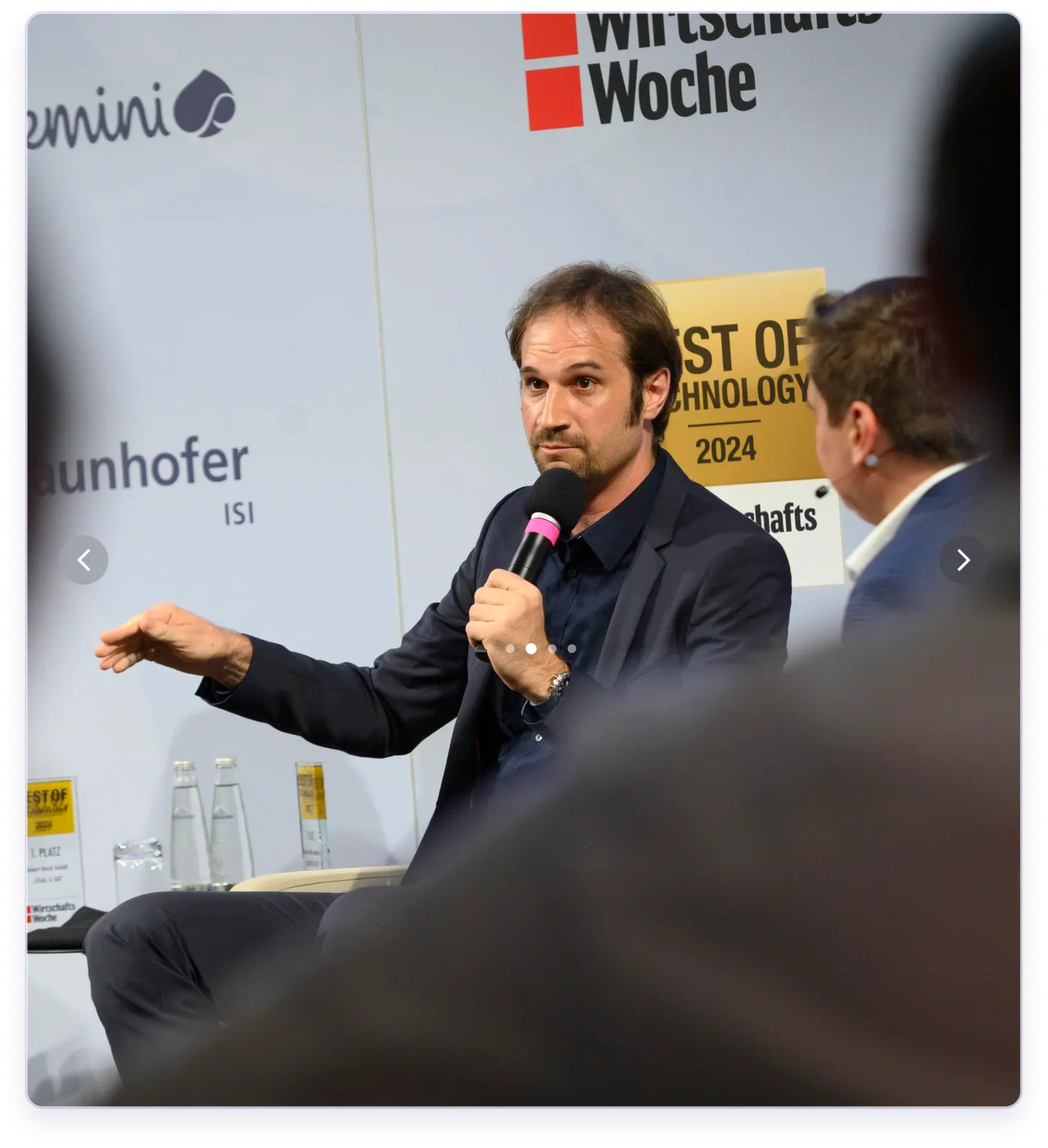 Ein Mann im Anzug spricht in ein Mikrofon während einer Podiumsdiskussion, neben ihm sitzt ein weiterer Mann.