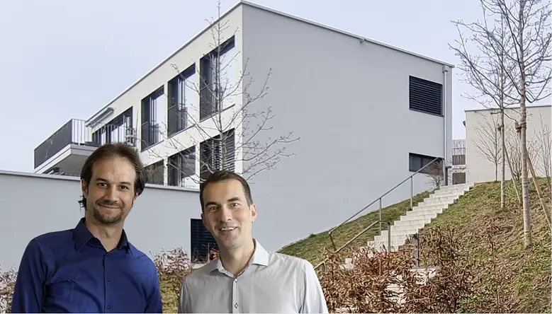 Zwei lächelnde Männer stehen vor einem modernen Gebäude mit Treppe und karger Landschaft im Hintergrund.