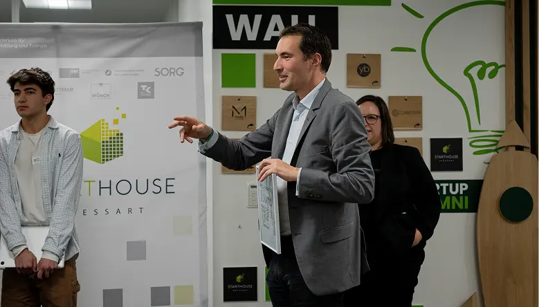 Mann im grauen Anzug hält eine Urkunde und gestikuliert, während zwei Personen im Hintergrund stehen in einem Raum mit Start-up-Dekoration.