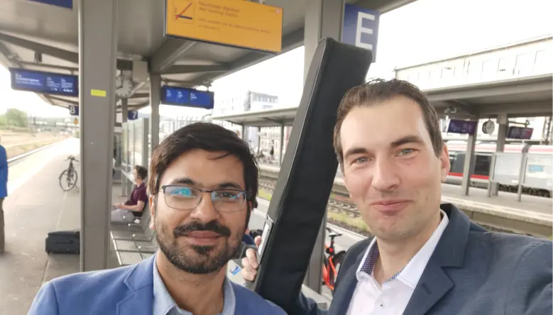 Zwei Männer in Anzügen posieren lächelnd am Bahnhof, einer hält eine Gitarrentasche.