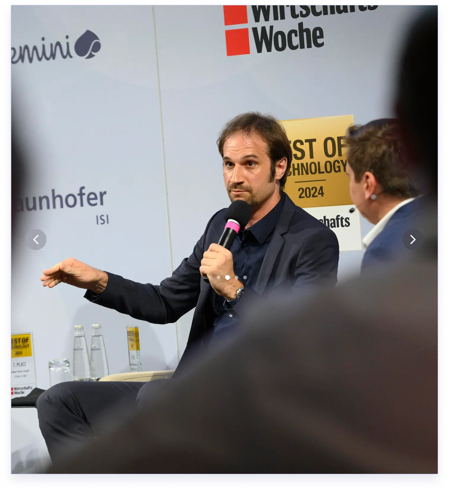 Mann im Anzug hält Mikrofon und spricht bei einer Podiumsdiskussion mit Publikum im Vordergrund.