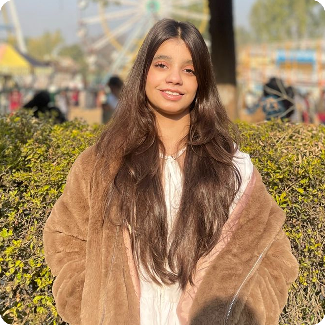 Young woman with long dark hair wearing a brown fur coat smiling outdoors with a Ferris wheel in the background.