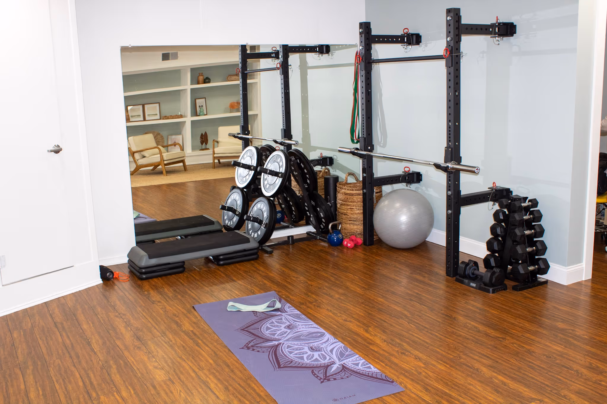 Home gym with a yoga mat, weight plates on a rack, dumbbells, exercise ball, and resistance bands against a wall with large mirrors. Reynolds Physical Therapy + Wellness