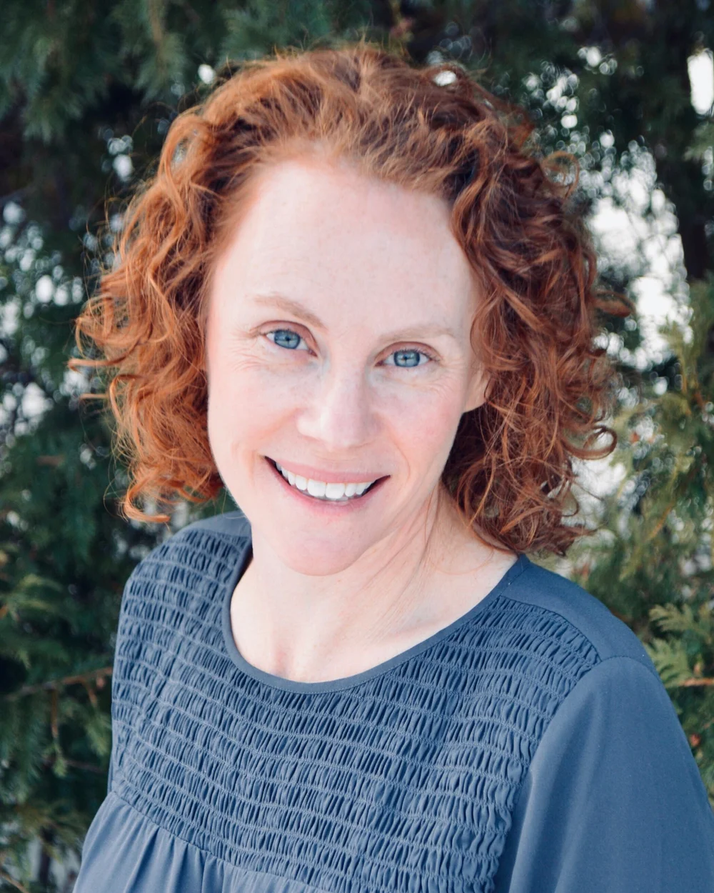 Smiling woman with curly red hair and blue eyes wearing a textured blue top outdoors with green trees in the background. Reynolds Physical Therapy + Wellness