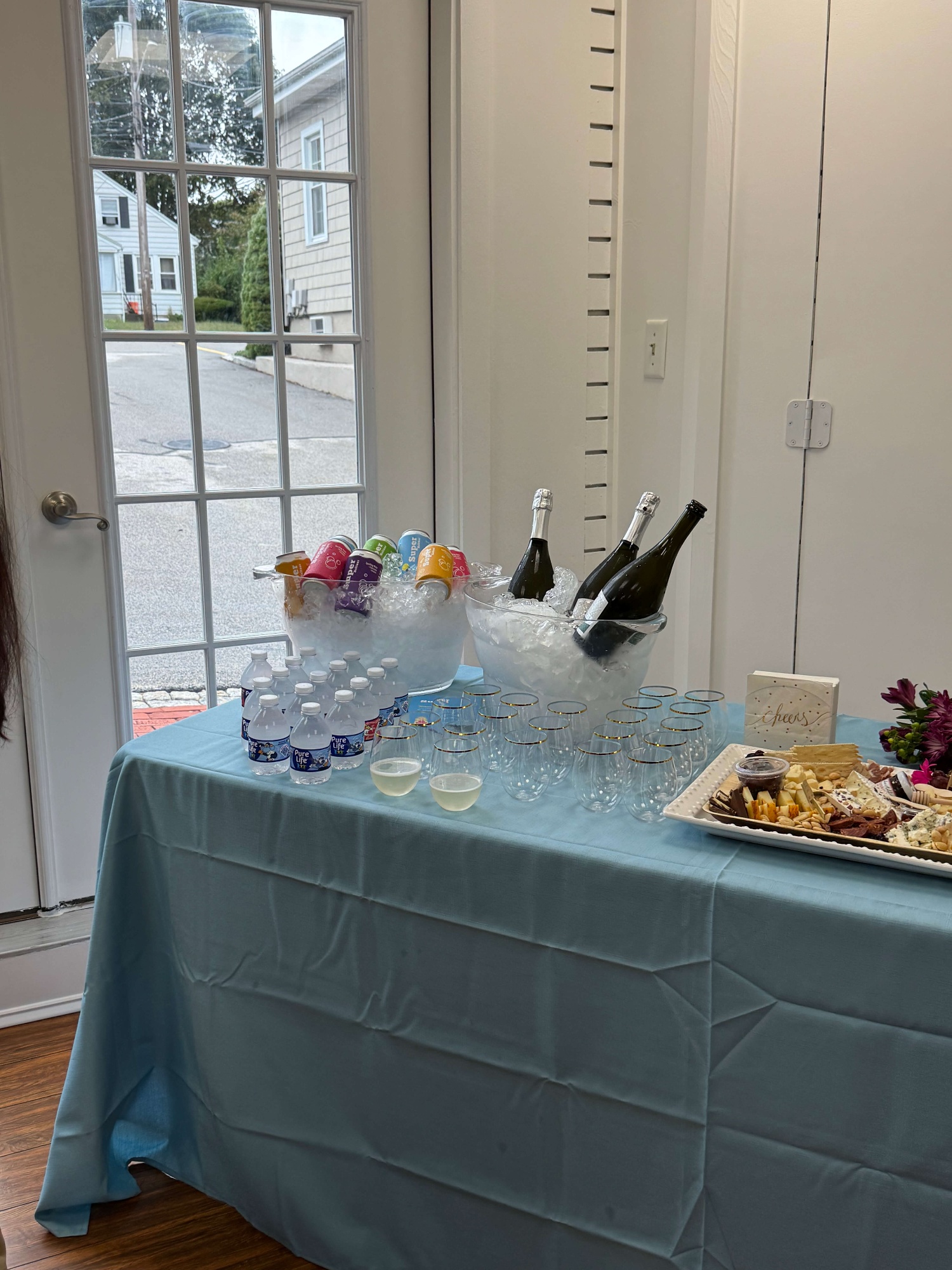 Table with blue tablecloth displaying bottles of sparkling wine and canned drinks in ice buckets, water bottles, empty glasses with gold rims, and a cheese and cracker platter next to a 'Cheers' napkin.